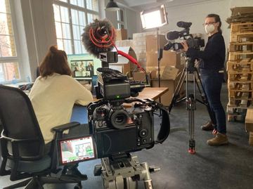 Kamerafrau mit Mundschutz filmt eine Interviewsituation in einem Lager